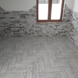 Anmir - Ułożona podłoga z płytek imitujących drewno w jodełkę oraz ściana wyłożona częściowo cegiełkami z fugą, widoczne niedokończone wykończenie ściany przy oknie.