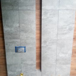 Anmir - Ściana w trakcie układania szarych płytek imitujących kamień, z widocznymi otworami na instalację sanitarną i niebieskim szablonem montażowym, sąsiedztwo ściany z panelami drewnopodobnymi.