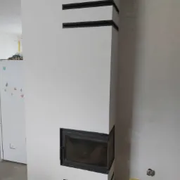 Biały kominek narożny z czarnymi akcentami w trakcie wykańczania, widoczne niedoskonałości na ścianie i podłodze, zarys lodówki w tle.