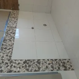 Jasne płytki podłogowe z dekoracyjnym pasem z mozaiki kamiennej w odcieniach szarości i bieli, widoczne otwory na instalacje podłogowe.