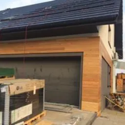 Fragment elewacji domu z garażem, wykończony drewnianą okładziną. Widoczne materiały budowlane w opakowaniach przed garażem i na paletach z boku. Dach pokryty ciemną dachówką.