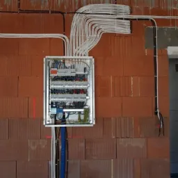 Otwarta skrzynka bezpiecznikowa z licznymi białymi kablami ułożonymi w równoległe wiązki, prowadzącymi w górę po ceglanej ścianie w surowym stanie.