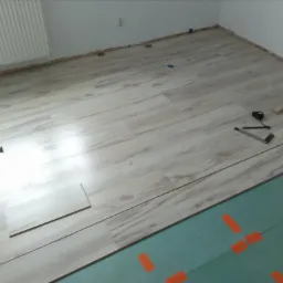 Proces układania paneli podłogowych w jasnym odcieniu drewna, widoczne narzędzia pomiarowe, młotek, podkład z oznaczeniami, oraz niedokończony fragment podłogi przy grzejniku.