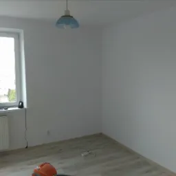 Pomalowane na biało wnętrze pokoju z drewnopodobną podłogą, grzejnikiem pod oknem i niebieską lampą wiszącą. Na podłodze leży przedłużacz i odkurzacz.