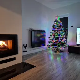 Salon z kominkiem i choinką świąteczną, szare panele podłogowe, telewizor z zimowym krajobrazem, czarny grzejnik.