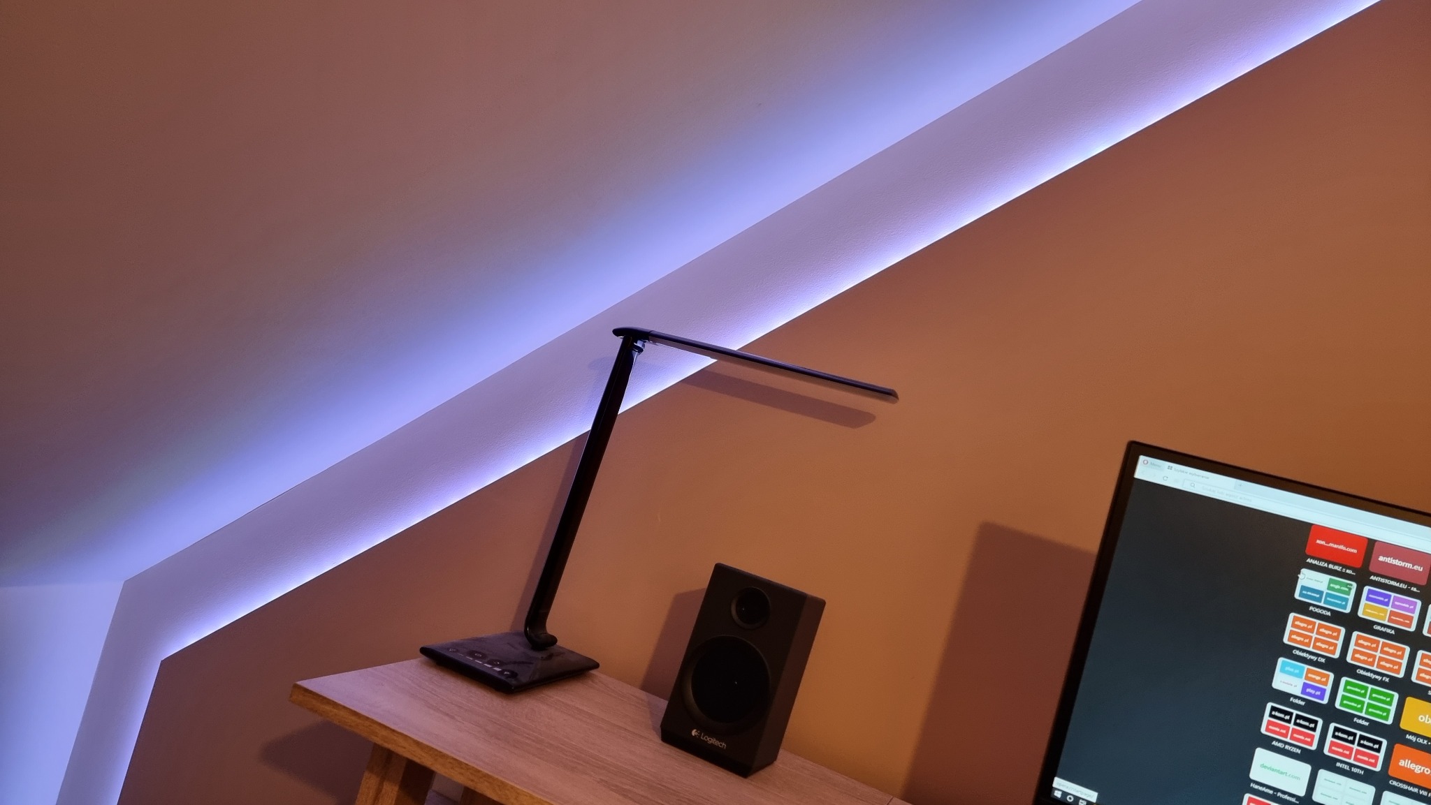 Oświetlenie LED wzdłuż krawędzi sufitu w skosie, biurko z lampką i głośnikiem Logitech, monitor z otwartymi oknami przeglądarki.