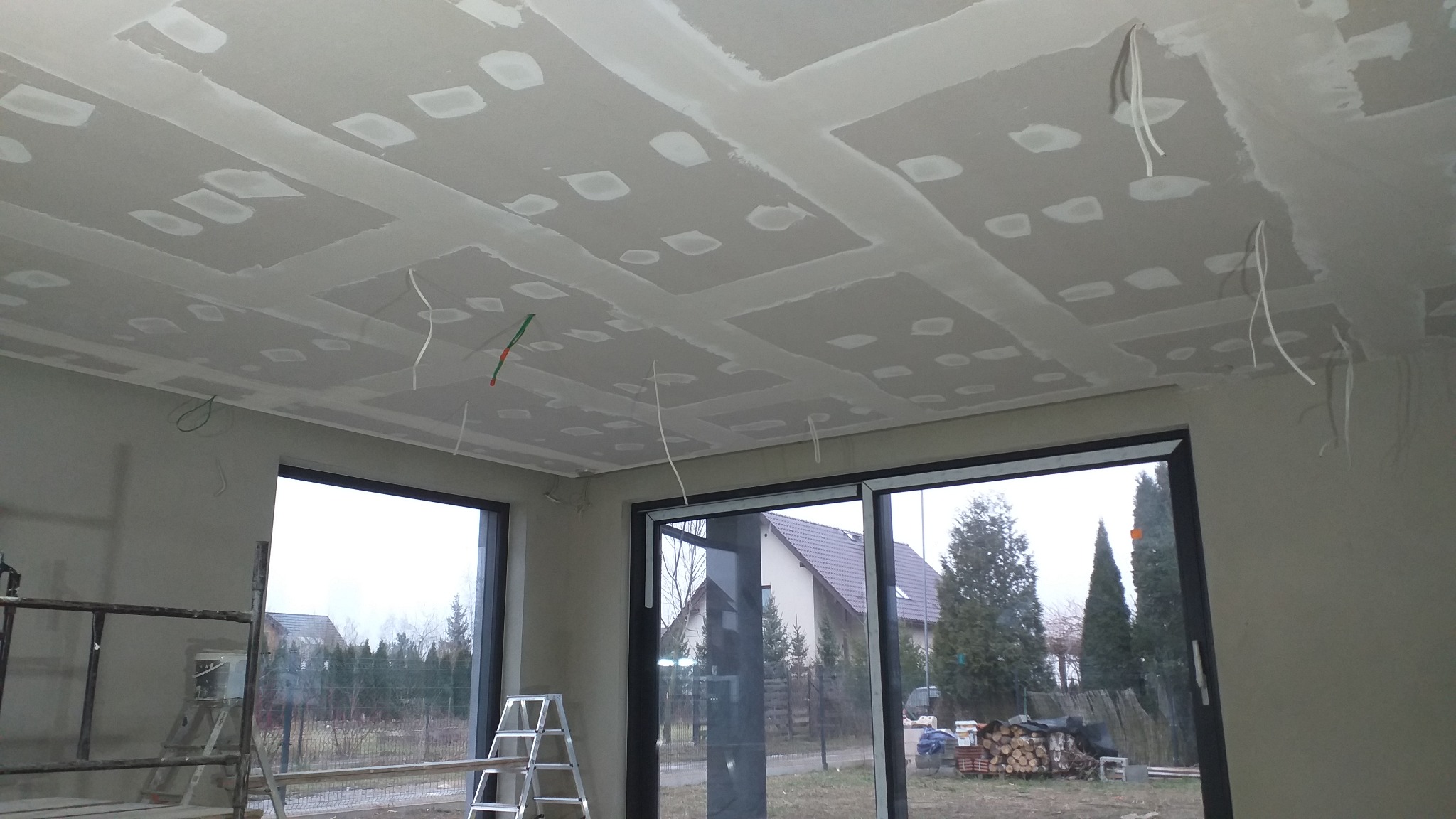 Sufit podwieszany z płyt gipsowo-kartonowych w trakcie montażu, widoczne szpachlowane łączenia, wystające przewody elektryczne, drabina aluminiowa i okno z widokiem na posesję.