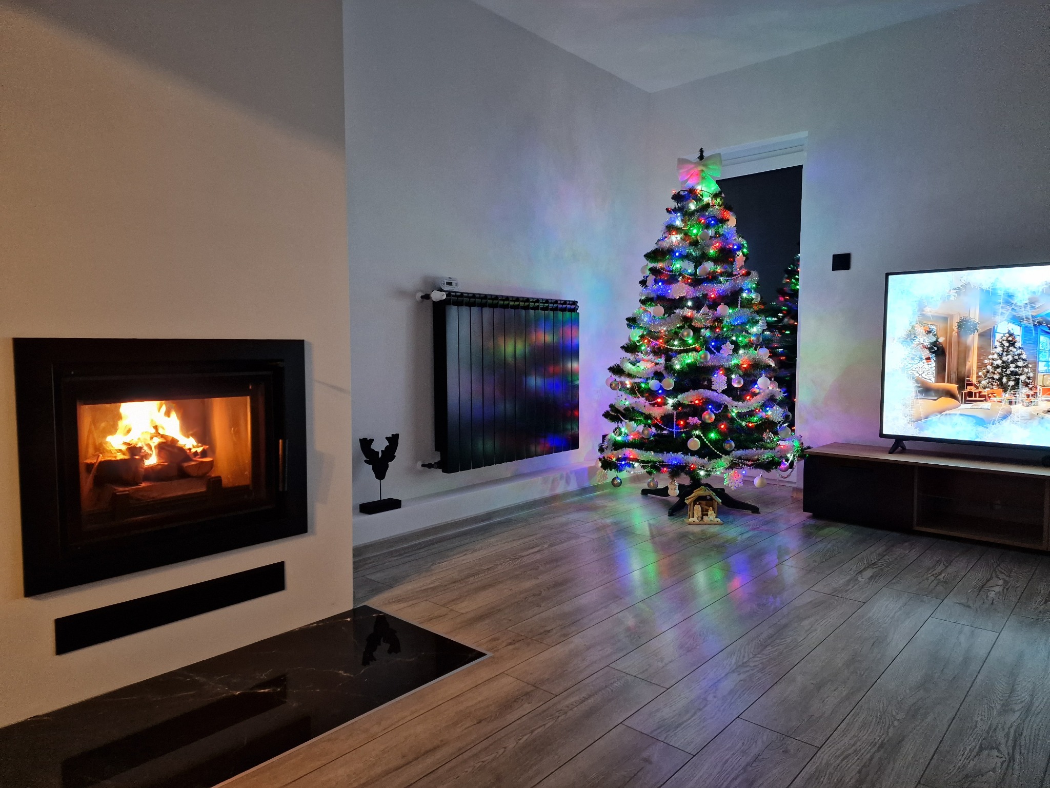 Salon z kominkiem i choinką świąteczną, szare panele podłogowe, telewizor z zimowym krajobrazem, czarny grzejnik.