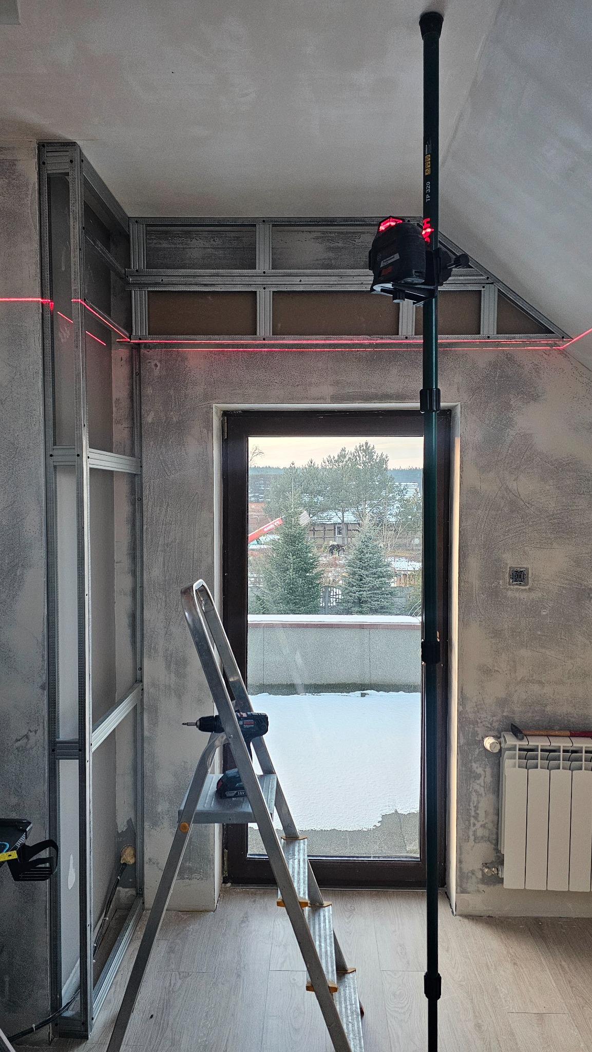 Montaż konstrukcji metalowej pod zabudowę w pomieszczeniu ze ścianami w stanie surowym, widoczny laser krzyżowy wyznaczający poziom, drabina z wkrętarką akumulatorową, za oknem widok na zimowy...