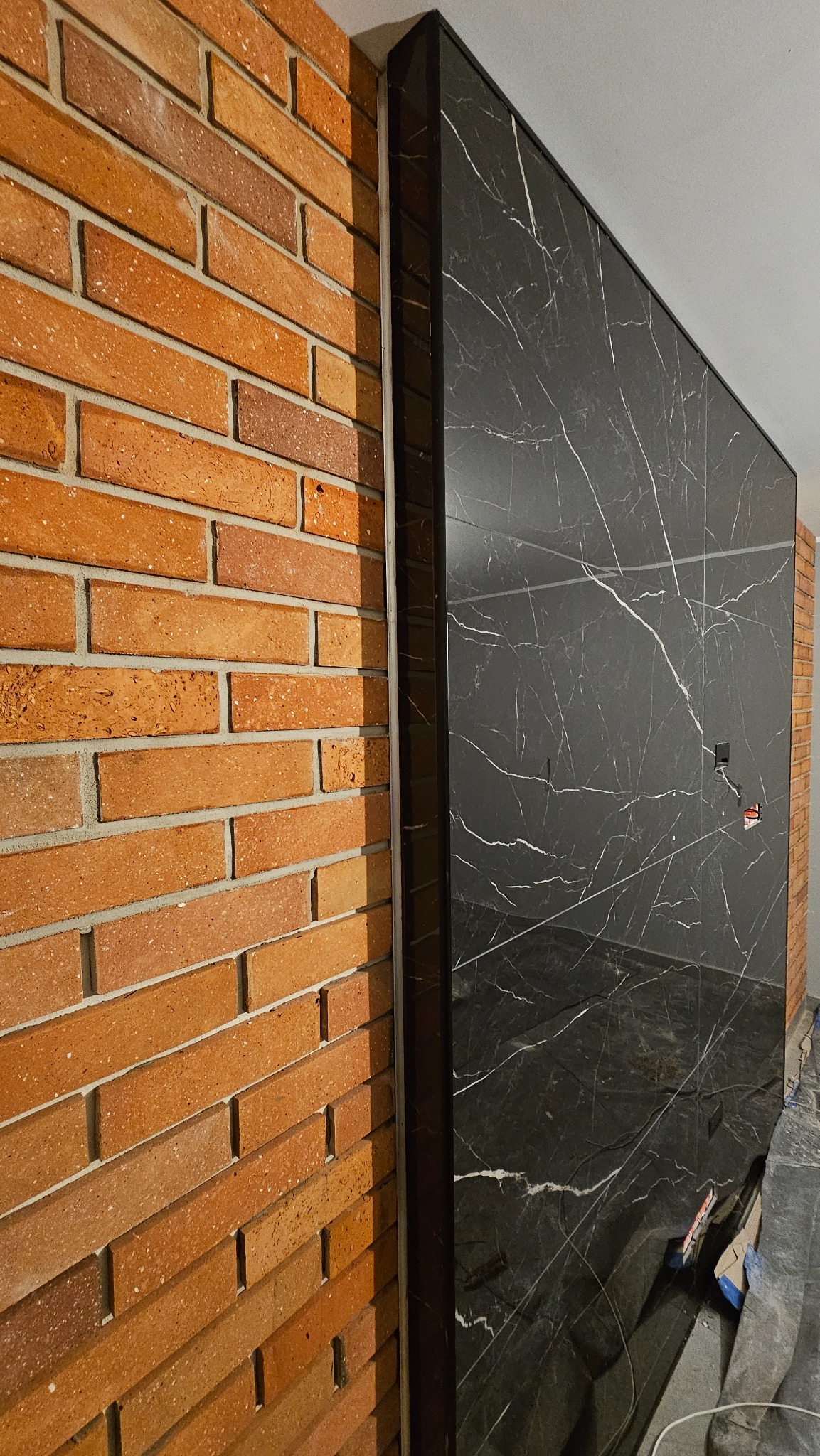 Ściana wykończona czerwoną cegłą sąsiadująca z panelem imitującym czarny marmur z białymi żyłami, obramowanym czarną listwą.