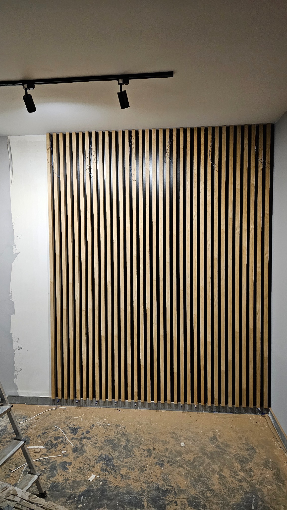 Montaż dekoracyjnych paneli lamelowych na ścianie, widoczne niedociągnięcia w wykończeniu ściany obok, przewody wystające zza paneli, surowy stan podłogi i drabina w rogu.