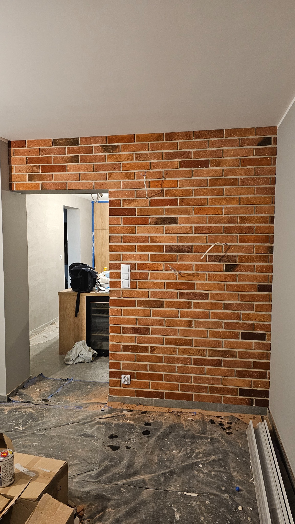 Ściana z cegły klinkierowej w trakcie wykańczania, widoczne puszki elektryczne, przewody i folia ochronna na podłodze, w tle widoczne elementy wyposażenia wnętrza.