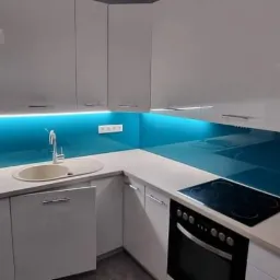 Nowoczesna kuchnia z błyszczącymi białymi szafkami i turkusowym panelem szklanym między blatem a szafkami wiszącymi, oświetlonym od dołu.