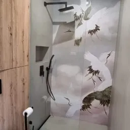 Nowoczesna kabina prysznicowa ze szklaną ścianką z motywem żurawi, czarna armatura, drewniane szafki w tle, widoczny fragment toalety.