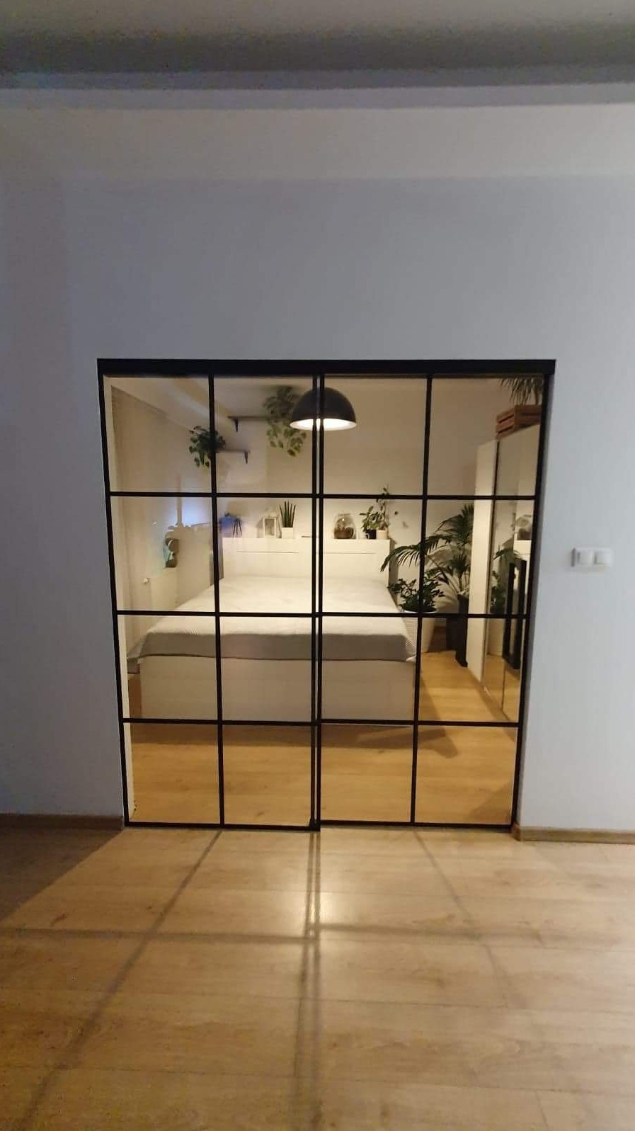 Dwuskrzydłowe drzwi loftowe przesuwne z czarnymi szprosami oddzielające sypialnię, widok na białe łóżko z roślinami i lampą wiszącą, podłoga drewniana.