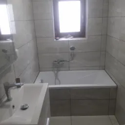 Wykończona łazienka z szarymi płytkami, białą wanną, umywalką i toaletą, okno z widocznym parapetem.