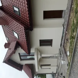 Nowo wybudowany dom jednorodzinny z czerwoną dachówką, jasną elewacją, widoczną drabiną i wiadrem farby przy wejściu, sugerujący świeże prace wykończeniowe.