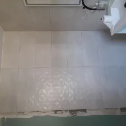 Ściana łazienki wyłożona jasnymi, kwadratowymi płytkami ceramicznymi z trójwymiarowym wzorem geometrycznym, widoczny fragment umywalki i niedokończone wykończenie dolnej części ściany.