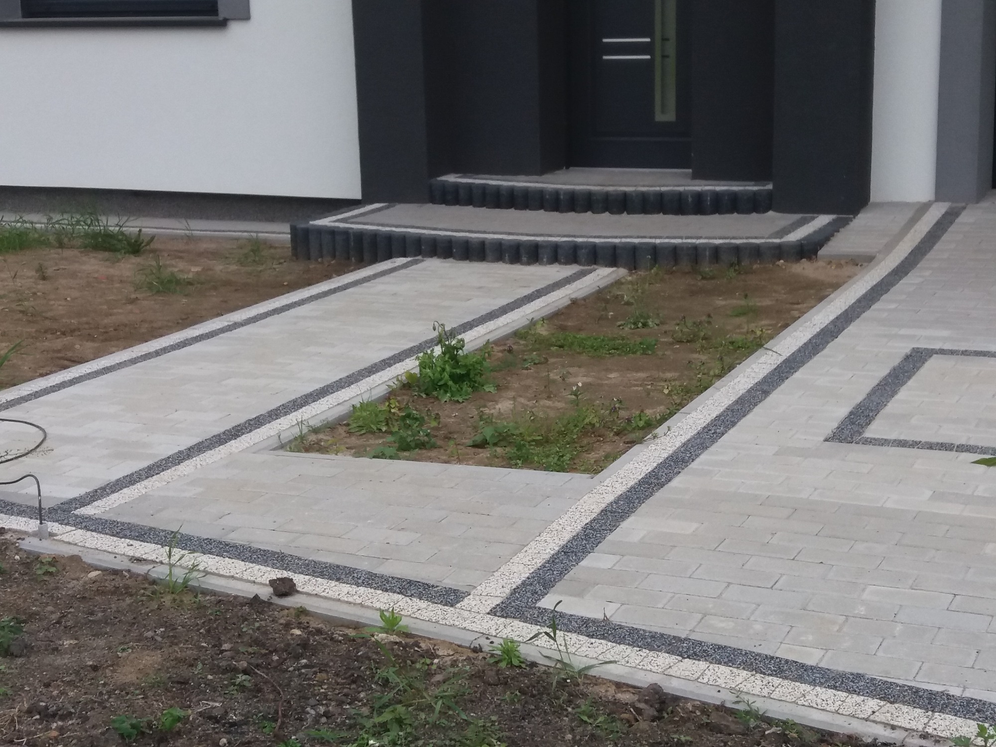 Nowoczesny podjazd z szarej kostki brukowej z dekoracyjnymi obramowaniami z drobnych kamieni, prowadzący do wejścia domu z ciemnymi drzwiami i schodami.