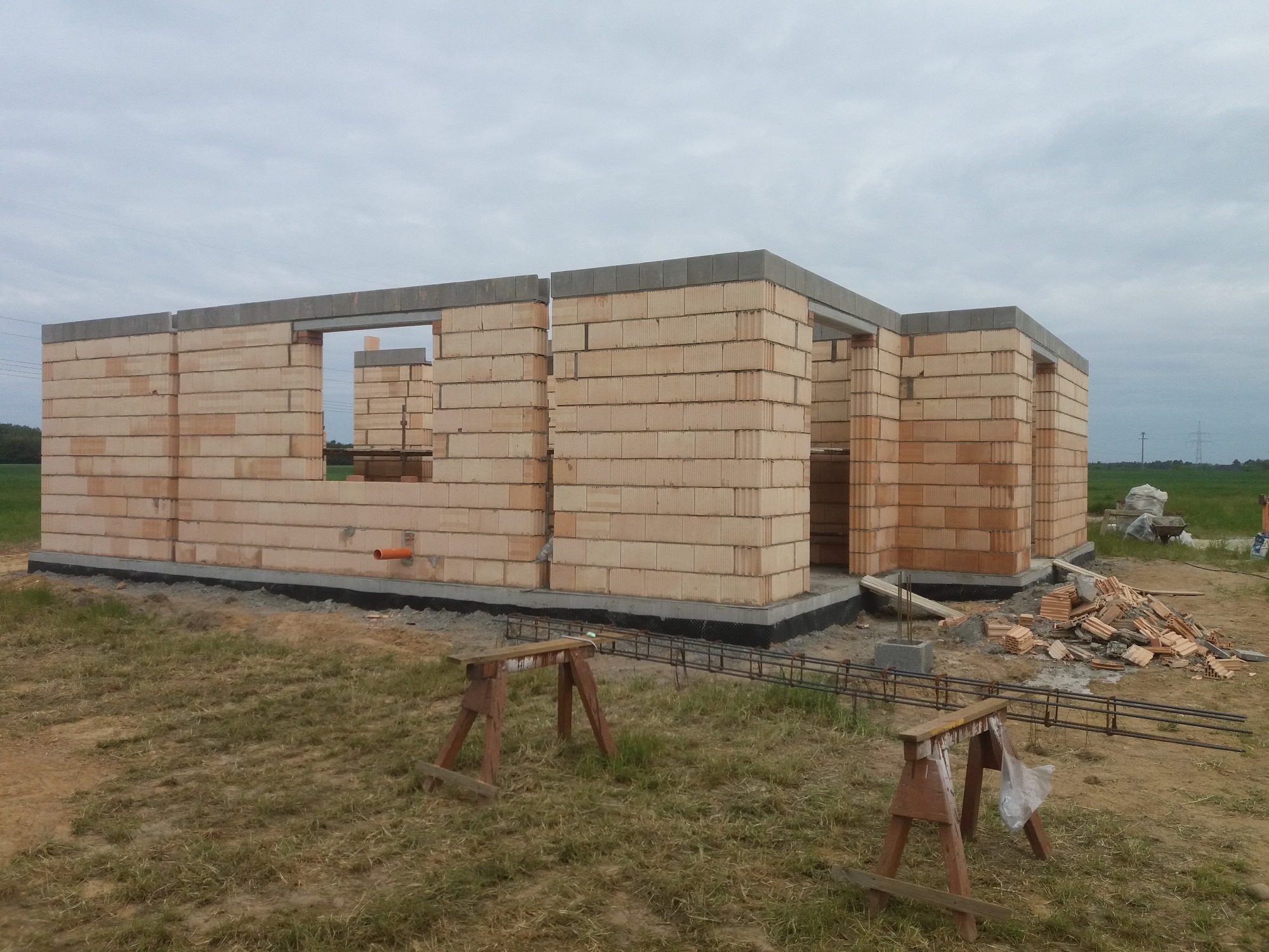 Wznoszenie ścian zewnętrznych budynku z bloczków ceramicznych, widoczne nadproża okienne i drzwiowe, teren budowy z drewnianymi kozłami i zbrojeniem.