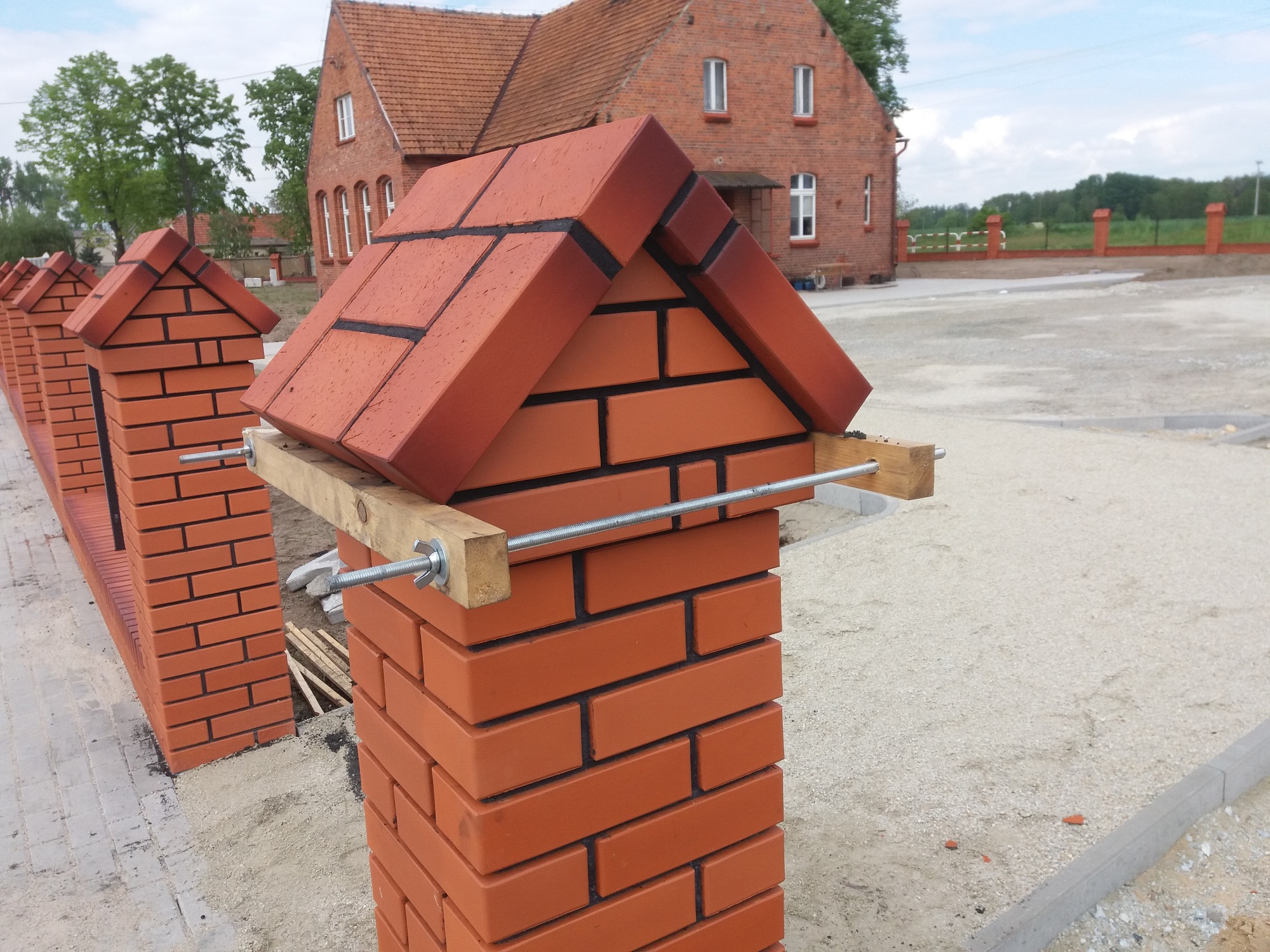 Budowa ogrodzenia z cegły klinkierowej, widoczne słupki z daszkami w trakcie wznoszenia, wsparte drewnianymi belkami i metalowymi śrubami, w tle budynek z czerwonej cegły.