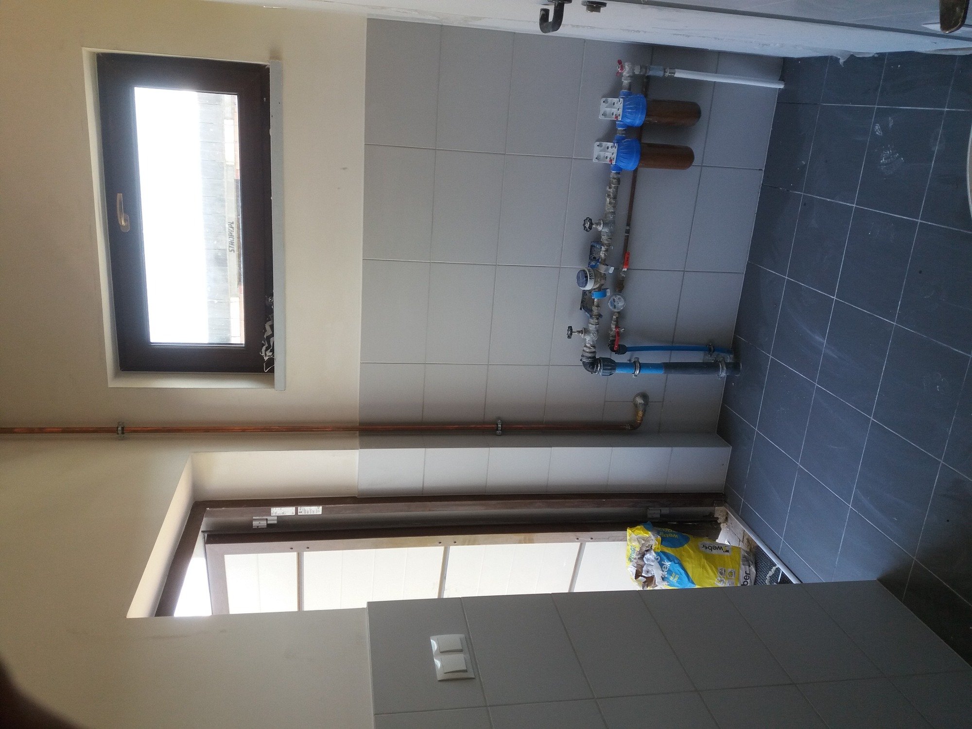 Wnętrze łazienki w trakcie remontu, widoczne płytki na ścianach i podłodze, instalacja wodna z filtrami, okno i drzwi, worek z materiałami budowlanymi w kącie.