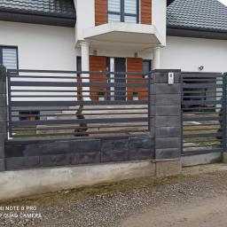 JM OPERATIVE - Nowoczesne ogrodzenie z szarego betonu łupanego i poziomych przęseł stalowych, z furtką i domofonem, na tle białego domu z elementami drewnianymi.