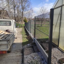 JM OPERATIVE - Nowoczesne ogrodzenie panelowe z podmurówką betonową, widok z boku z perspektywy niskiej, obok zaparkowany samochód dostawczy z paletami, w tle drzewa bez liści i błękitne niebo.