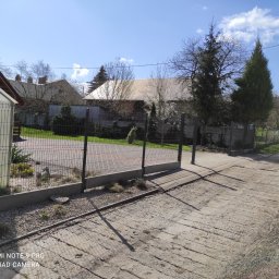 JM OPERATIVE - Widok na posesję z panelowym ogrodzeniem na betonowej podmurówce, z furtką i podjazdem z betonowych płyt, w tle zabudowania gospodarcze i drzewa.