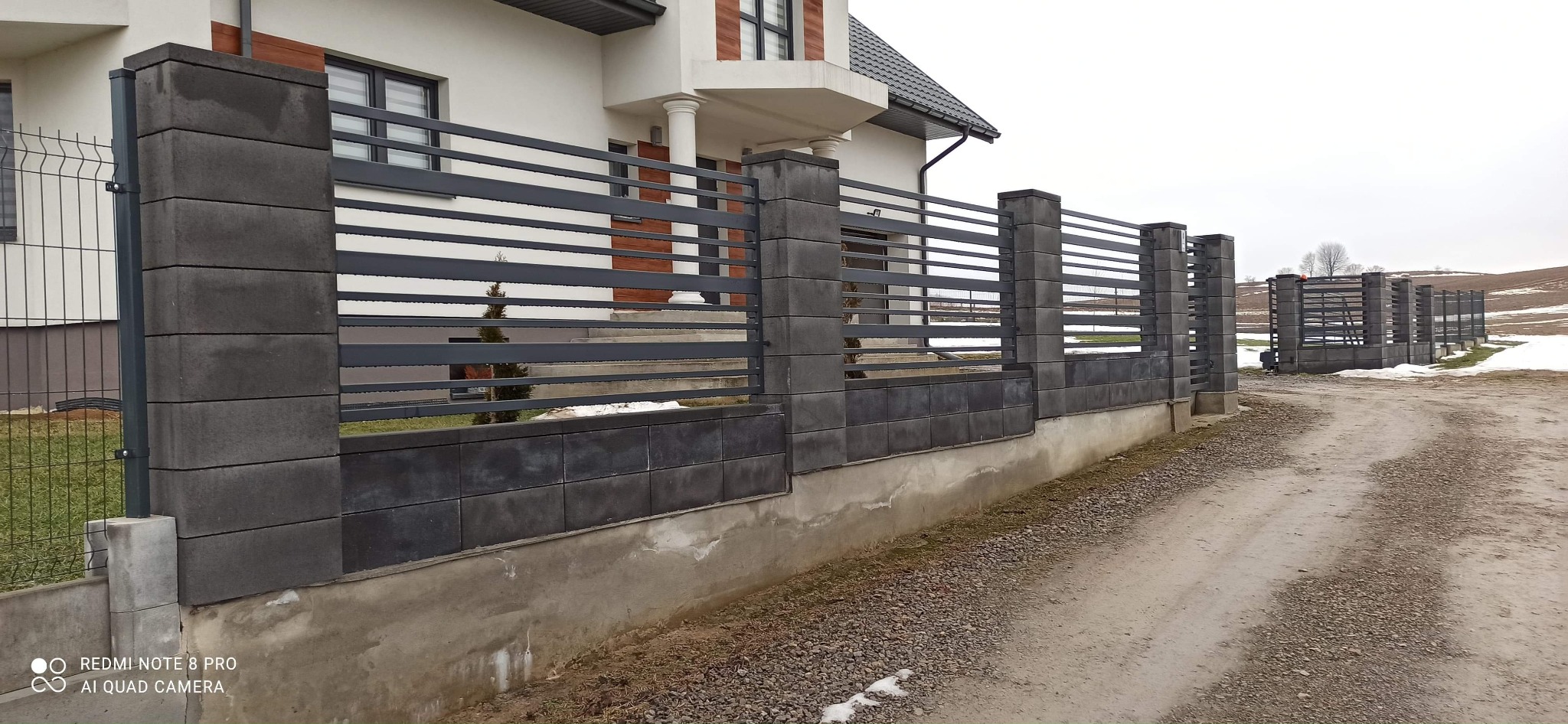 Nowoczesne ogrodzenie z betonowych bloczków i poziomych, stalowych przęseł, widok na dom z białymi kolumnami i częściowo ośnieżony teren w tle.