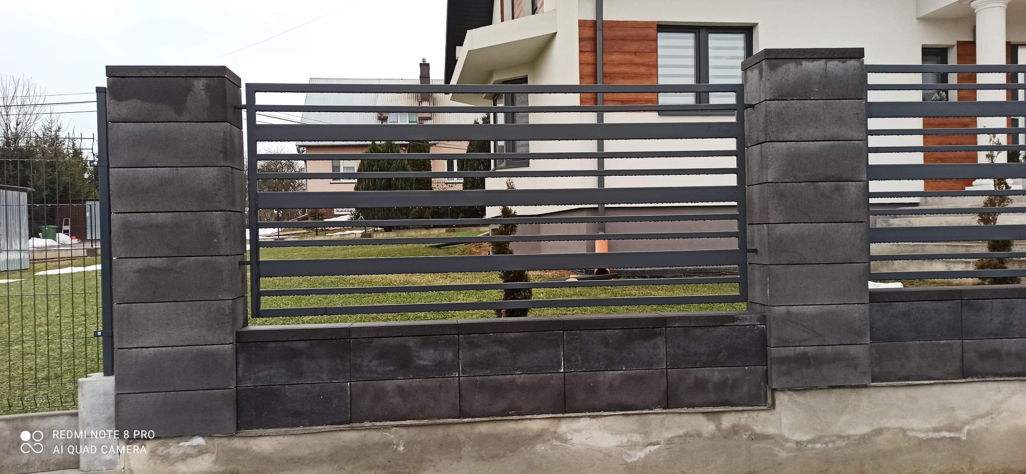 Nowoczesne ogrodzenie z poziomych, szarych paneli metalowych i słupków z szarej kostki betonowej, na tle trawnika i zabudowań.