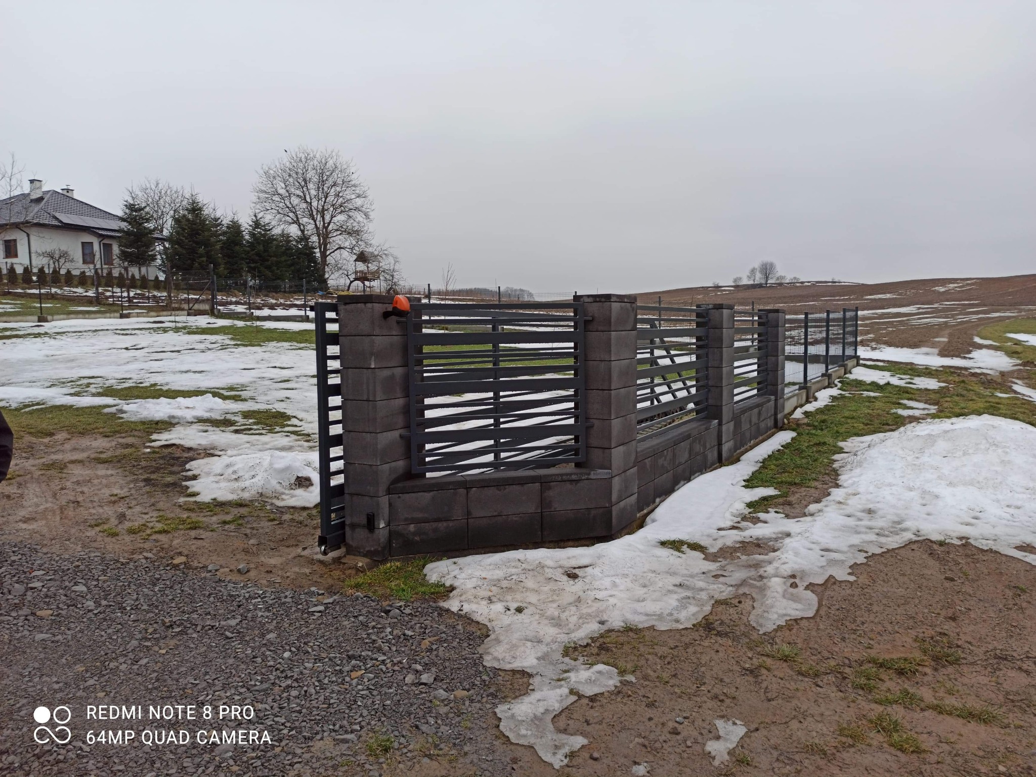 Nowoczesne ogrodzenie z szarego betonu modułowego i czarnych paneli metalowych, widok na zaśnieżone pole w pochmurny dzień.