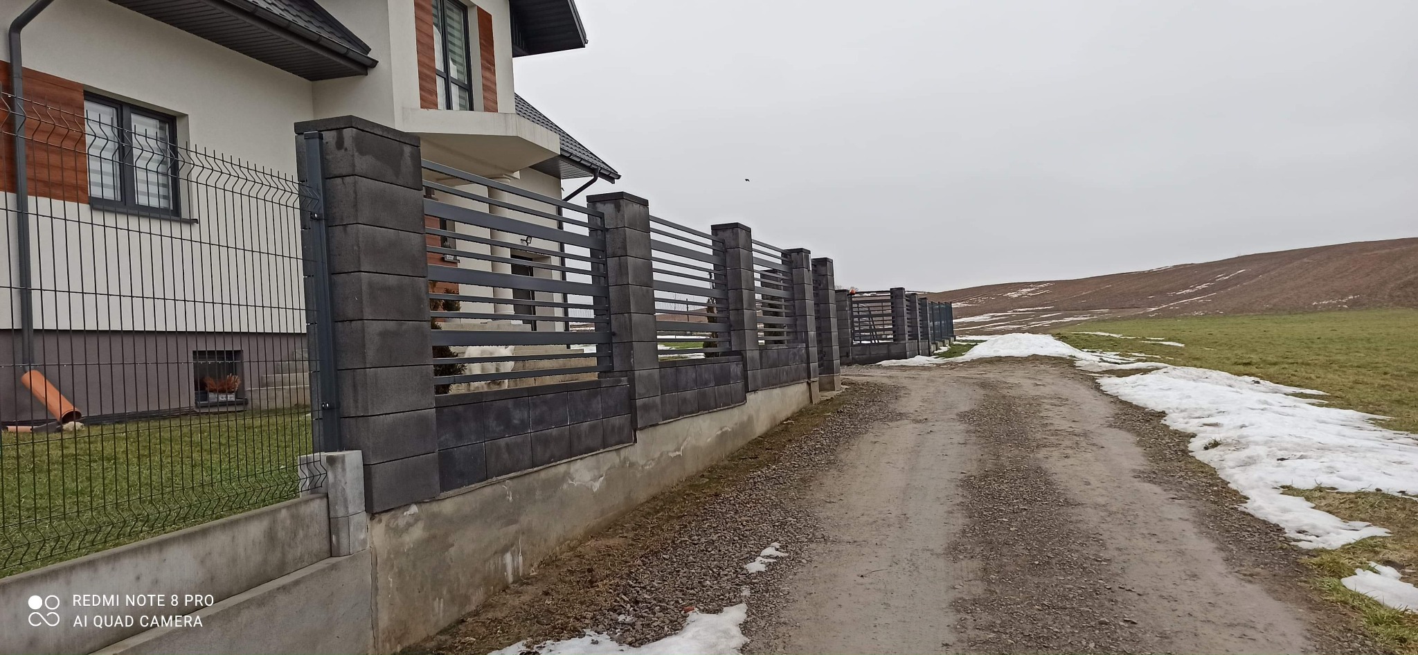 Nowoczesne ogrodzenie panelowe z betonowymi słupkami i podmurówką, widok z polnej drogi w pochmurny dzień, częściowo pokryte śniegiem.