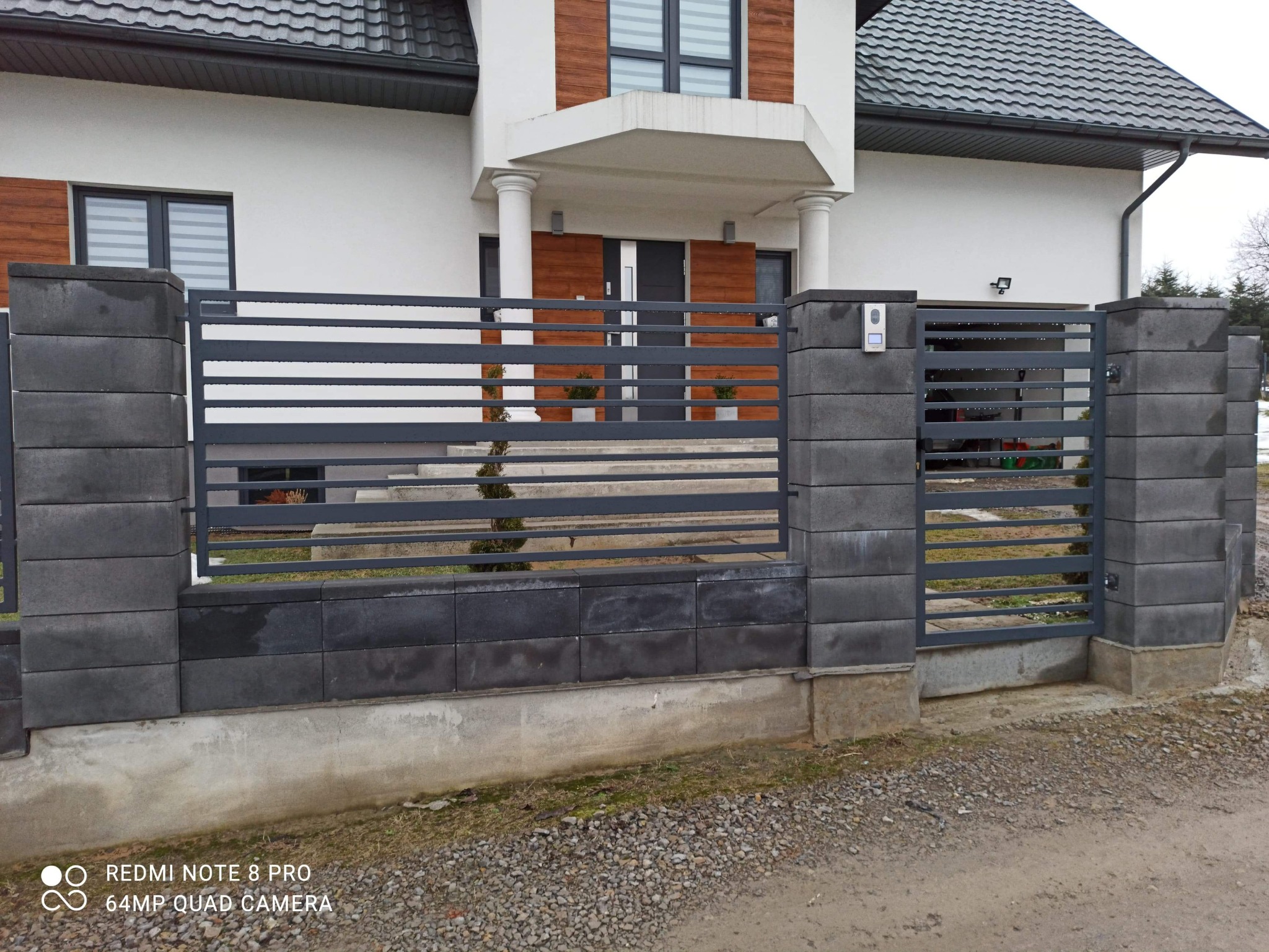 Nowoczesne ogrodzenie z szarego betonu łupanego i poziomych przęseł stalowych, z furtką i domofonem, na tle białego domu z elementami drewnianymi.