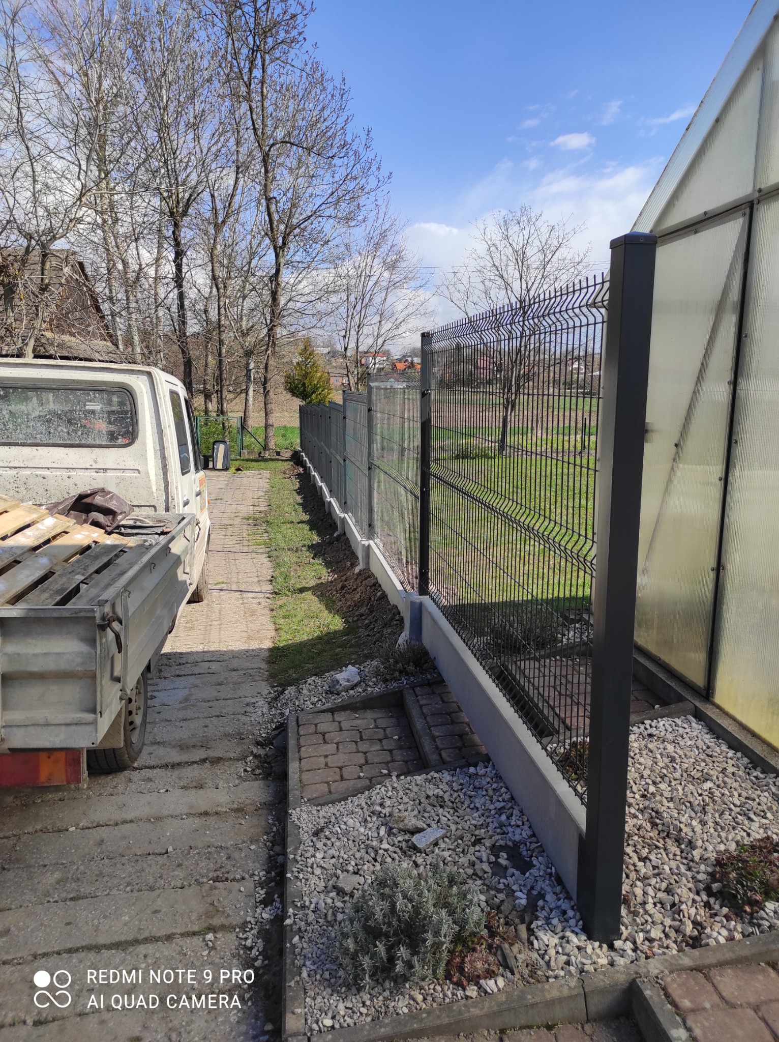 Nowoczesne ogrodzenie panelowe z podmurówką betonową, widok z boku z perspektywy niskiej, obok zaparkowany samochód dostawczy z paletami, w tle drzewa bez liści i błękitne niebo.