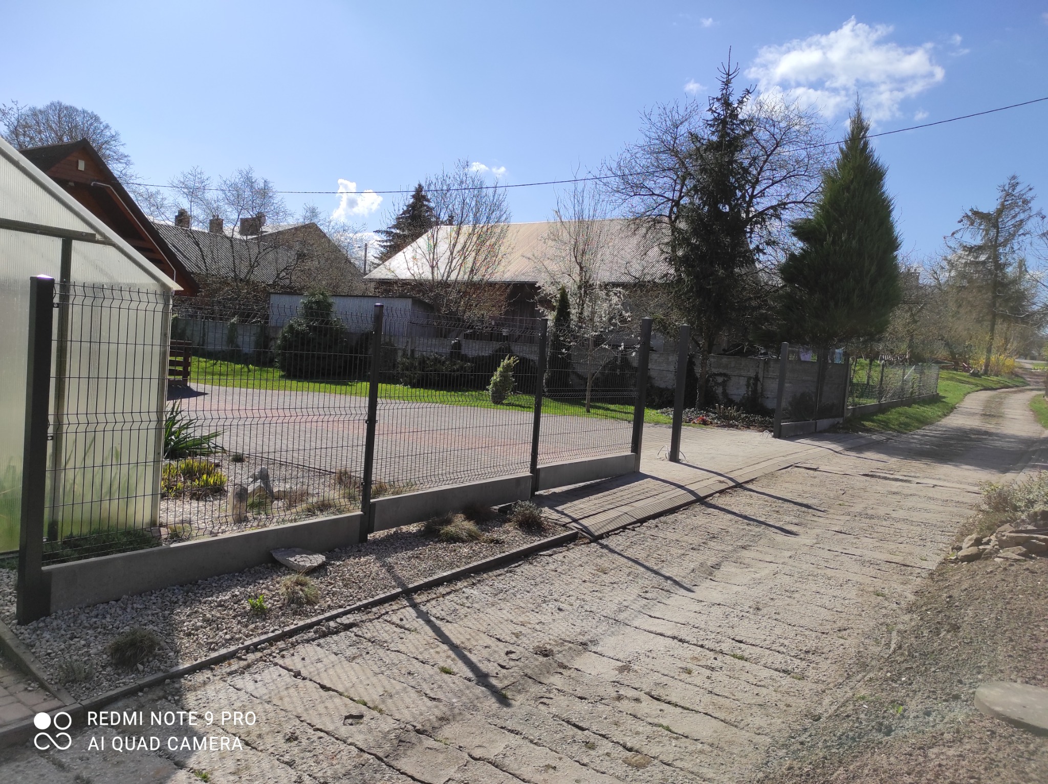 Widok na posesję z panelowym ogrodzeniem na betonowej podmurówce, z furtką i podjazdem z betonowych płyt, w tle zabudowania gospodarcze i drzewa.