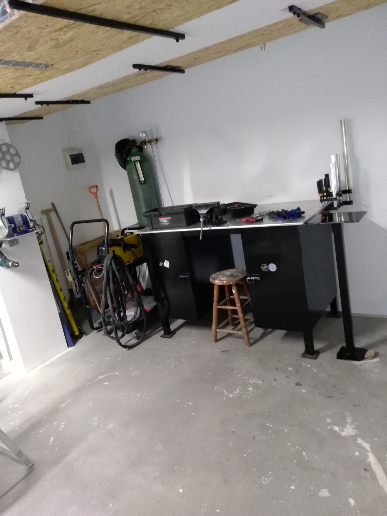 Warsztat: Czarny stół roboczy z narzędziami, butla z gazem i krzesło w pomieszczeniu z betonową podłogą i surowym stropem.