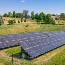 Rozległa farma fotowoltaiczna na łące, widok z góry, w tle wiejskie zabudowania i drzewa.