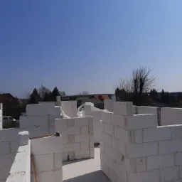 Wznoszenie ścian budynku z białych bloczków betonu komórkowego, widoczne zbrojenie i częściowo zapakowane bloczki, w tle zabudowa jednorodzinna i błękitne niebo.