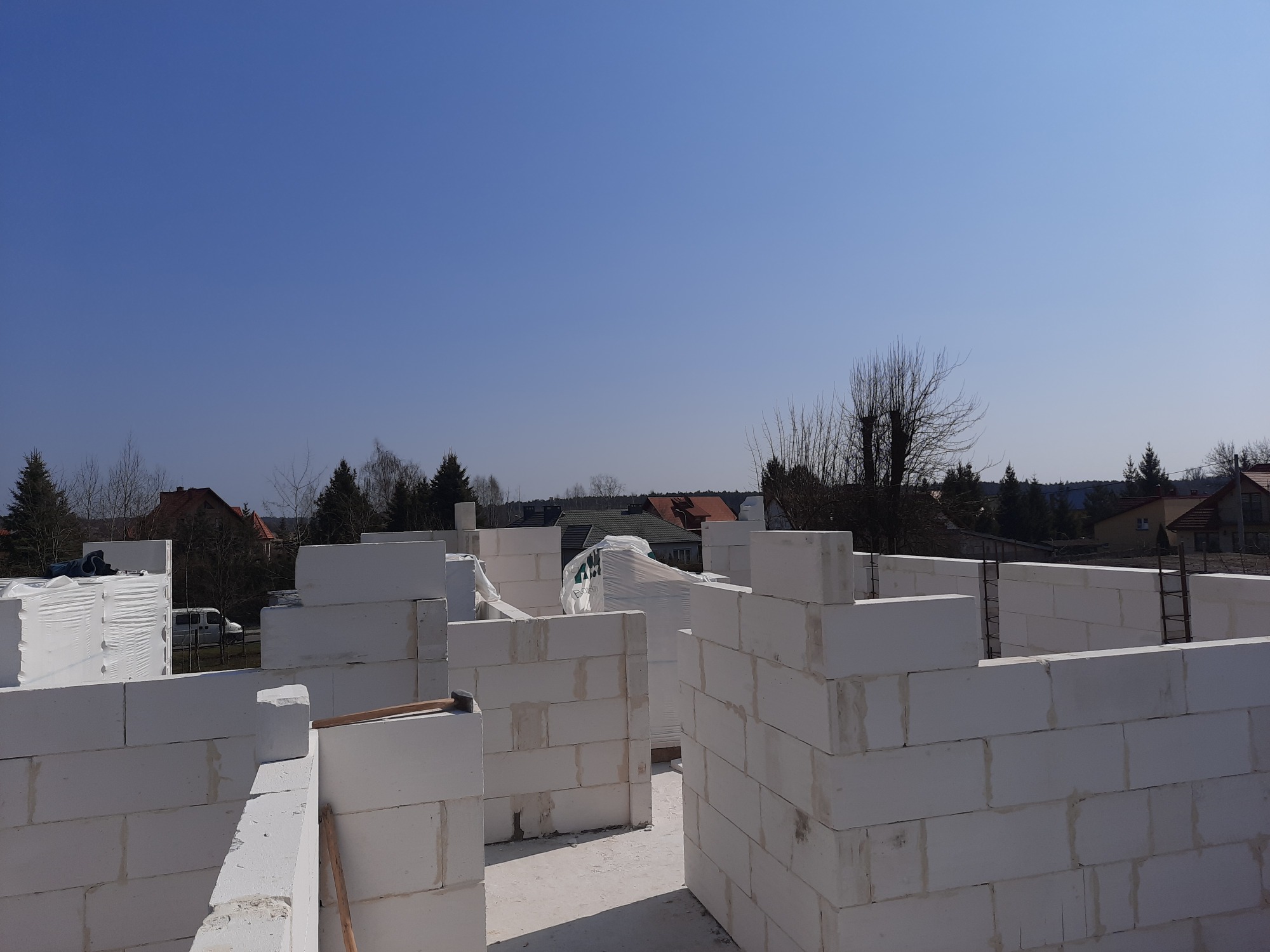 Wznoszenie ścian budynku z białych bloczków betonu komórkowego, widoczne zbrojenie i częściowo zapakowane bloczki, w tle zabudowa jednorodzinna i błękitne niebo.