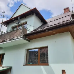 Ocieplony dom jednorodzinny z balkonem, brązowym dachem z blachy i nową elewacją w odcieniach bieli i zieleni, widoczne anteny satelitarne i naziemne.