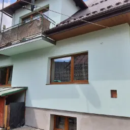 Dom jednorodzinny z jasną elewacją, brązowymi oknami i dachem, widok z boku, z fragmentem balkonu i przylegającą drewnianą szopą.