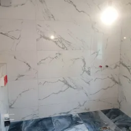 Ukończona łazienka z marmurowymi płytkami na ścianach i podłodze w odcieniach bieli i szarości, widoczne przyłącza wodne i oświetlenie sufitowe.