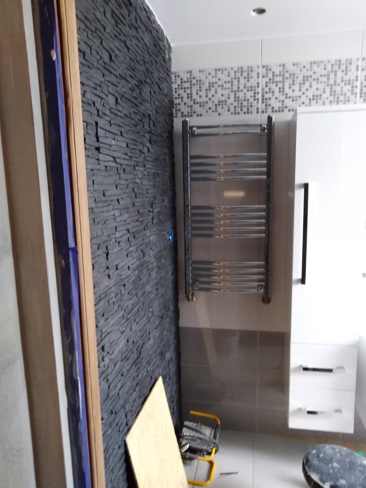 Ściana w łazience wykończona czarnym kamieniem dekoracyjnym, obok grzejnik drabinkowy, biała szafka z szufladami, widoczne narzędzia i płyta budowlana na podłodze.