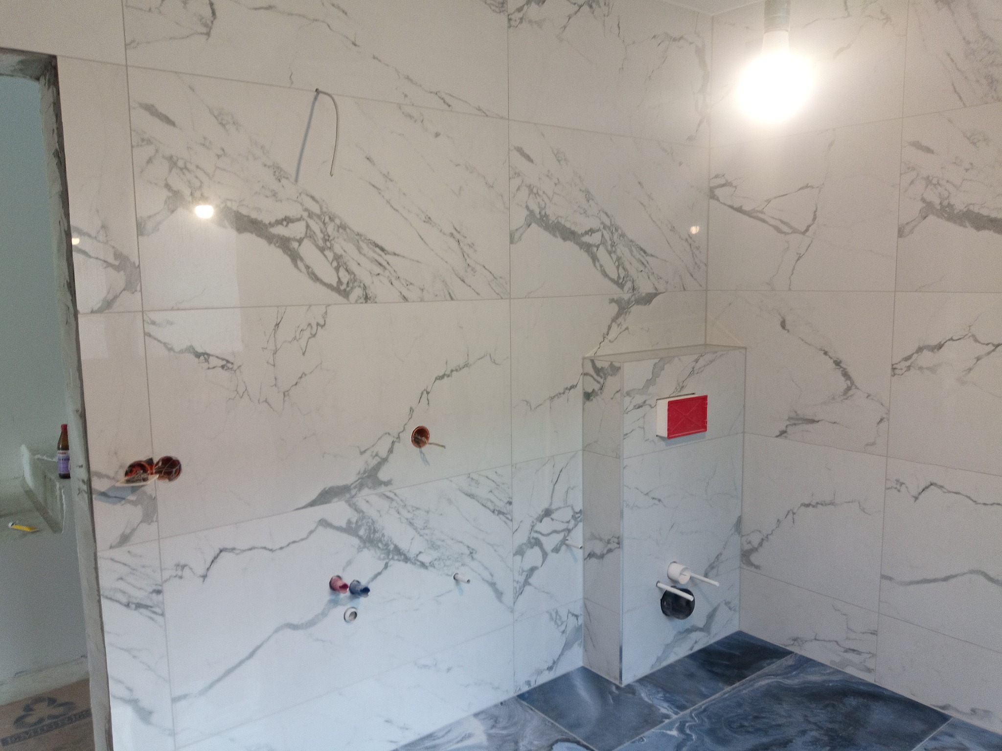 Łazienka w trakcie wykańczania z marmurowymi płytkami na ścianach i podłodze, widoczne wystające rury i instalacja podtynkowego WC.