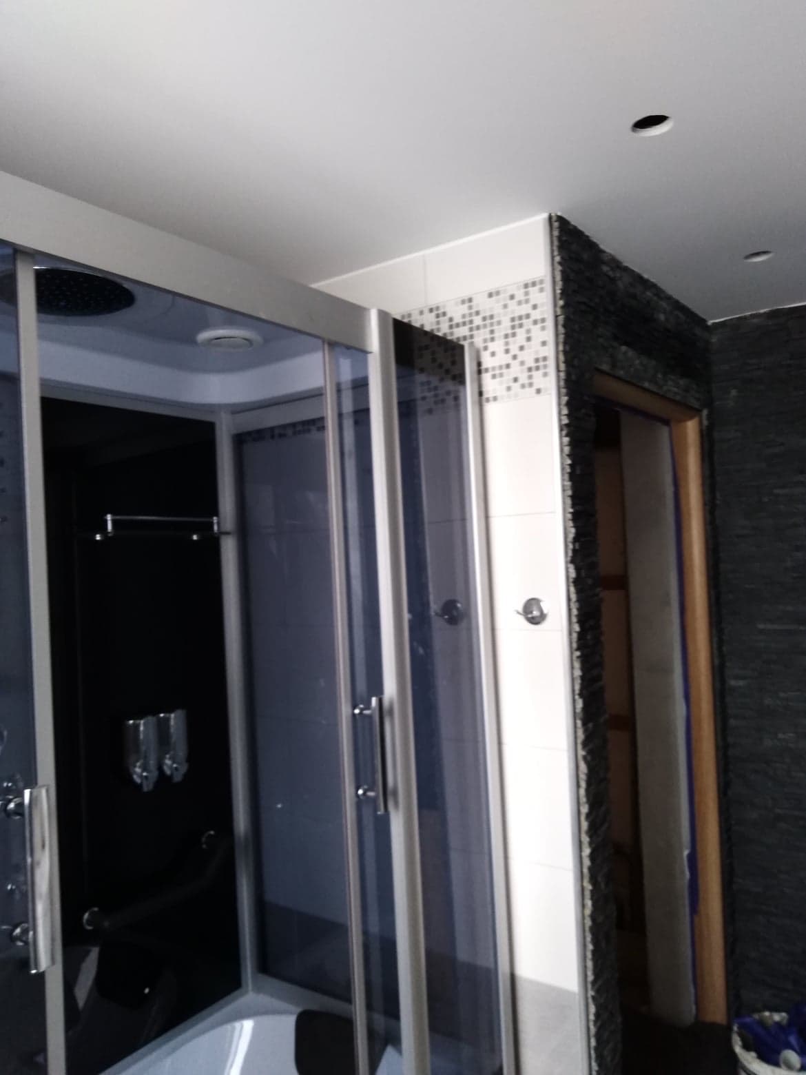 Czarna kabina prysznicowa z deszczownicą, chromowane uchwyty i dozowniki, białe płytki z mozaiką i czarna ściana z kamienia w wykończonej łazience.