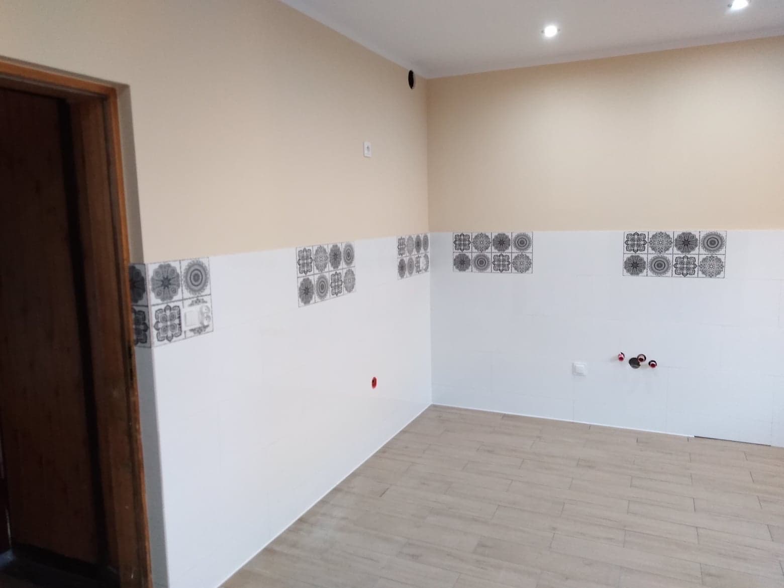 Świeżo wyremontowana kuchnia z białymi płytkami dekoracyjnymi na ścianach i panelami podłogowymi imitującymi drewno, widoczne instalacje hydrauliczne.