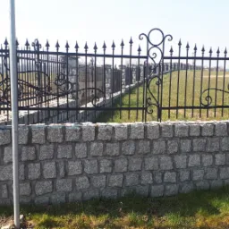 Granitowe ogrodzenie z czarnymi, metalowymi przęsłami o dekoracyjnym wzorze, widok na łąkę w tle.