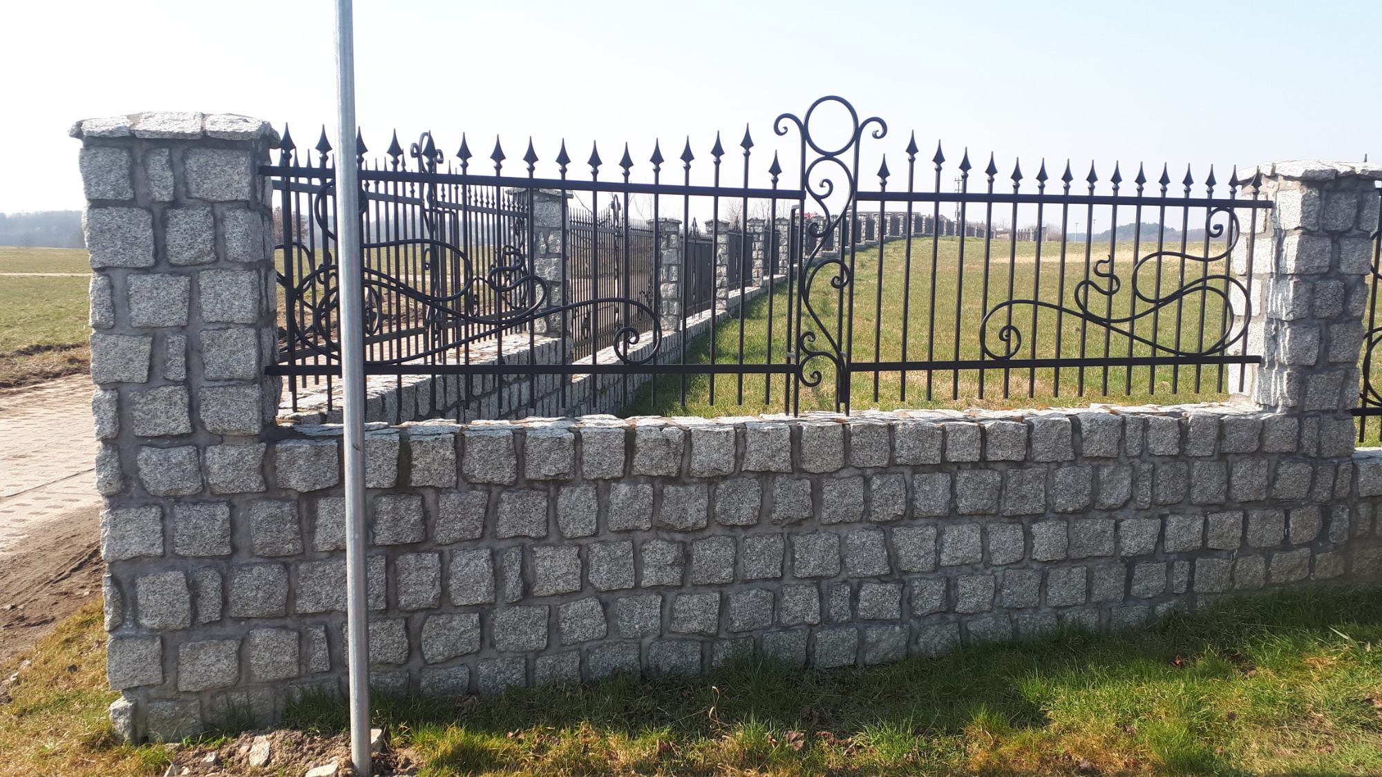 Granitowe ogrodzenie z czarnymi, metalowymi przęsłami o dekoracyjnym wzorze, widok na łąkę w tle.