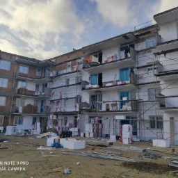 Wielopiętrowy budynek w trakcie prac związanych z dociepleniem, widoczne rusztowania, styropian w opakowaniach i fragmenty nieocieplonej ściany z cegły, pracownicy na rusztowaniach, ziemia...